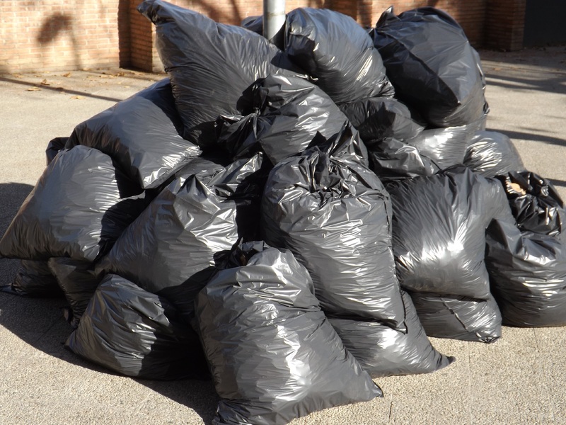 A large pile of black trash bags sits on pavement next to a brick building.