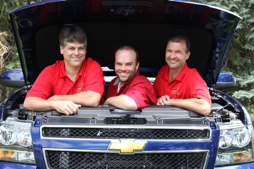 Three smiling men in red shirts pose from under the open hood of a blue Chevrolet truck.
