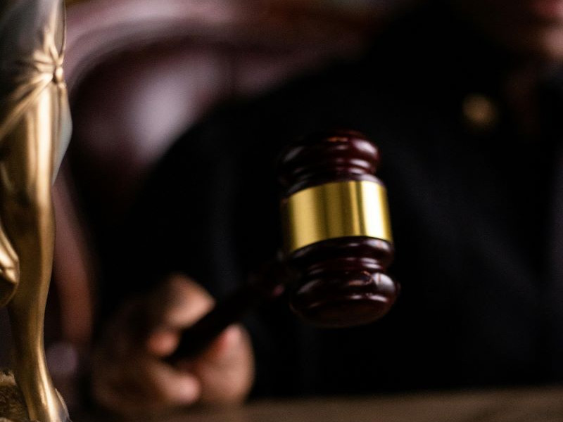 A hand holds a wooden gavel with a gold band next to a blurred golden statue of Lady Justice in a dark courtroom setting.