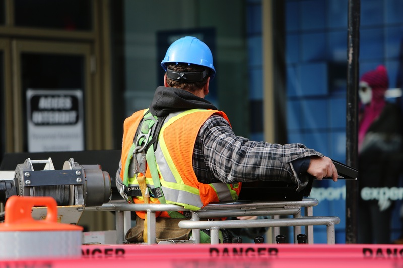 Construction worker in blue hard hat and orange vest operates equipment on a raised platform.