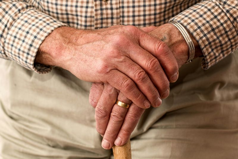 Close-up of an older person's hands, wearing a plaid shirt and gold ring, resting on a wooden cane.