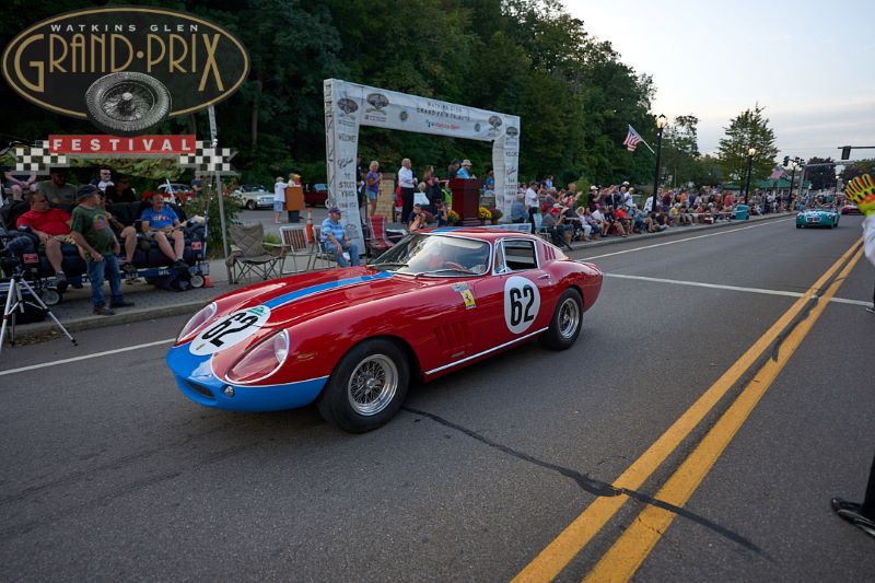 A red vintage race car with blue stripes and #62 drives past crowds at the Watkins Glen Grand Prix Festival.