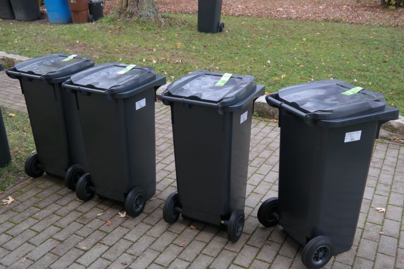 Four dark gray wheeled trash bins with green tags line a brick path next to a grassy lawn.