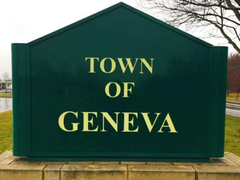 A dark green, peaked sign with 'TOWN OF GENEVA' in gold letters, set against a cloudy sky and bare trees.