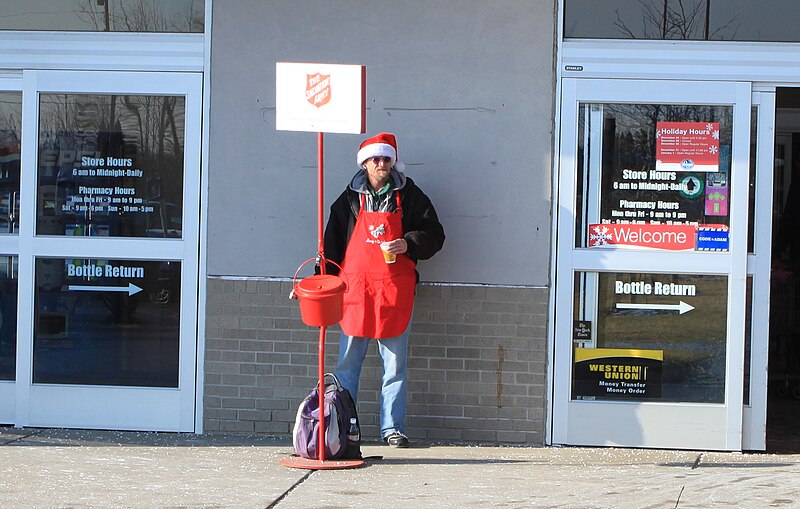 Man in Santa hat and red apron stands by a Salvation Army red kettle outside a Walmart entrance.