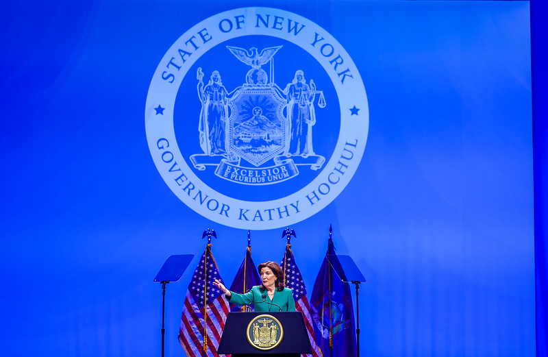 Governor Kathy Hochul speaks at a podium with the NY state seal against a blue backdrop with flags.