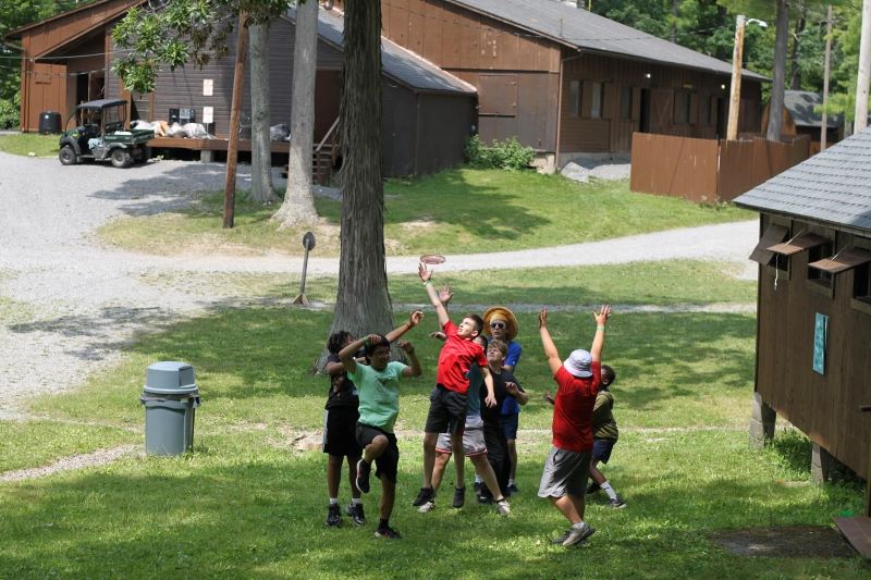 Children and an adult play frisbee on a grassy field at a summer camp with rustic brown cabins in the background.