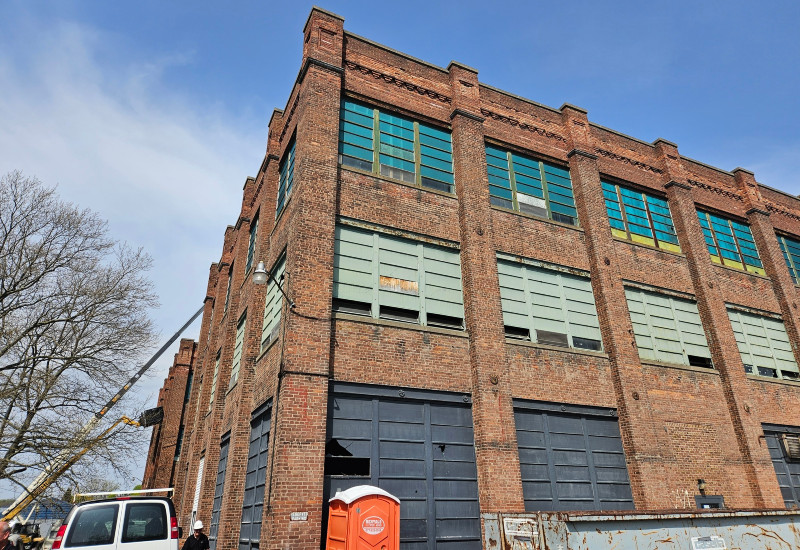 Red brick industrial building with large windows, some boarded, and ground-level doors. Construction equipment and a portable toilet are visible.