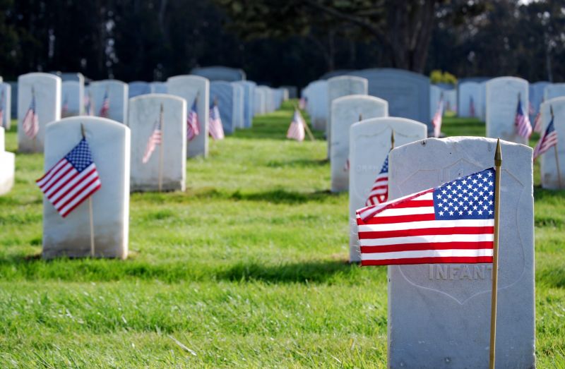 Rows of white headstones with small American flags on a green lawn in a military cemetery.