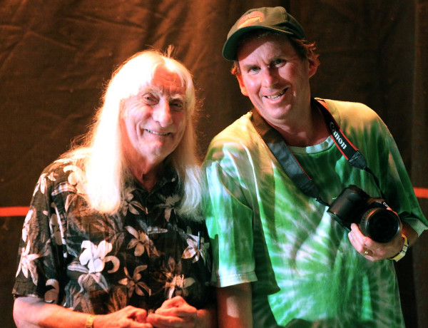 Greg Cotterill, wearing a green tie-dye shirt and cap, smiles with another man with long white hair.