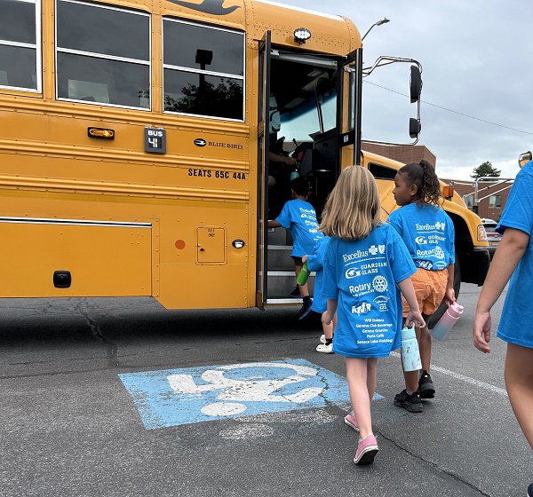 Children in matching blue shirts board a yellow school bus in a parking lot.