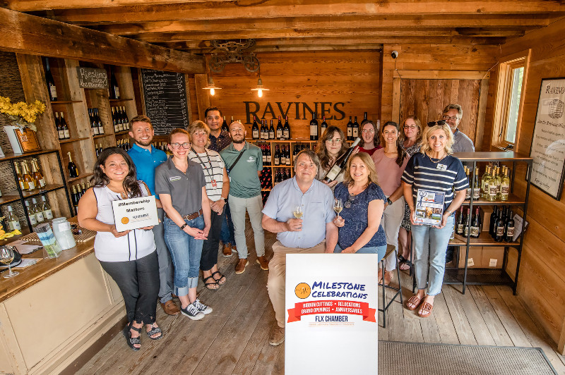 Group poses inside Ravines Wine Cellars with a "Milestone Celebrations FLX Chamber" sign and wine bottles.