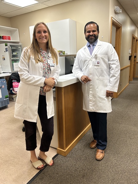A woman and a man in white lab coats stand smiling behind a counter in a medical office.