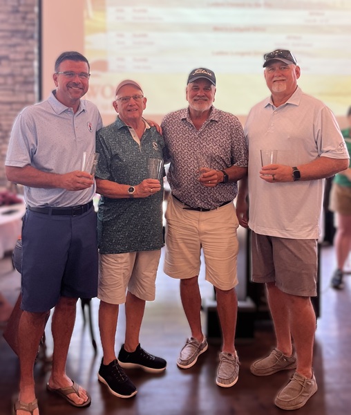 Four men in polo shirts and shorts hold drinks indoors at a casual event after a golf outing.