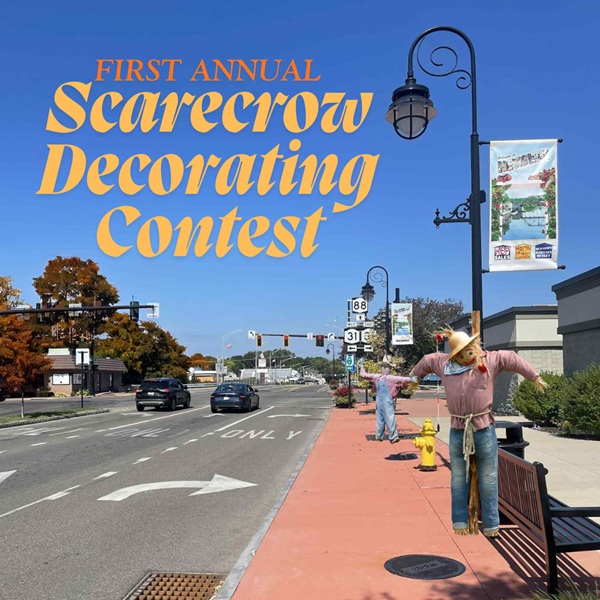 Scarecrows line a sunny street in Newark, NY, with a lamppost, street signs, and a banner for a decorating contest.