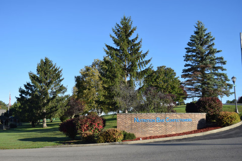 Brick sign for Nutrition Bar Confectioners at a facility entrance, surrounded by trees and landscaping under a blue sky.