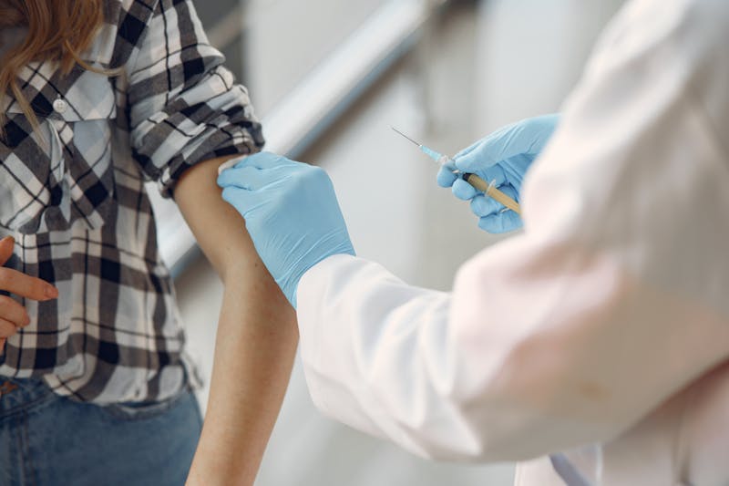 A gloved medical professional administers an injection to a patient's arm, who is wearing a plaid shirt.