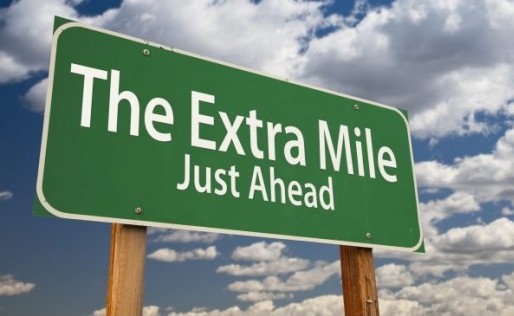 Green road sign reading "The Extra Mile Just Ahead" against a cloudy blue sky.