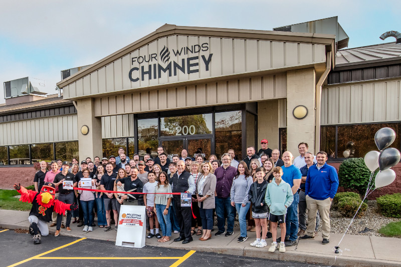 A large group, including a mascot, cuts a red ribbon in front of the Four Winds Chimney building.