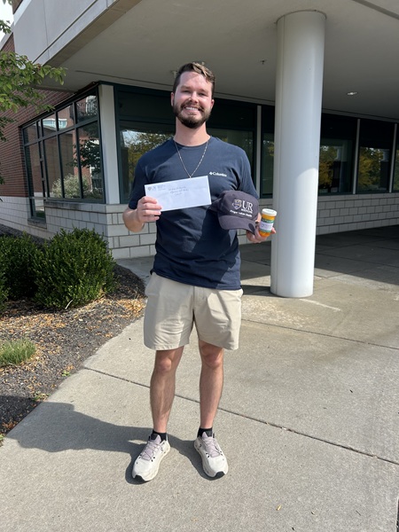 Man smiles, holding an envelope, pill bottle, and University of Rochester hat outside a building.
