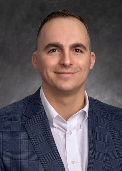 Headshot of a smiling man in a blue plaid jacket and white shirt against a gray background.