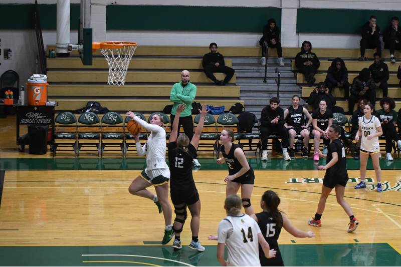 A Keuka College player in white and green shoots a layup against a Morrisville defender in black during a basketball game.