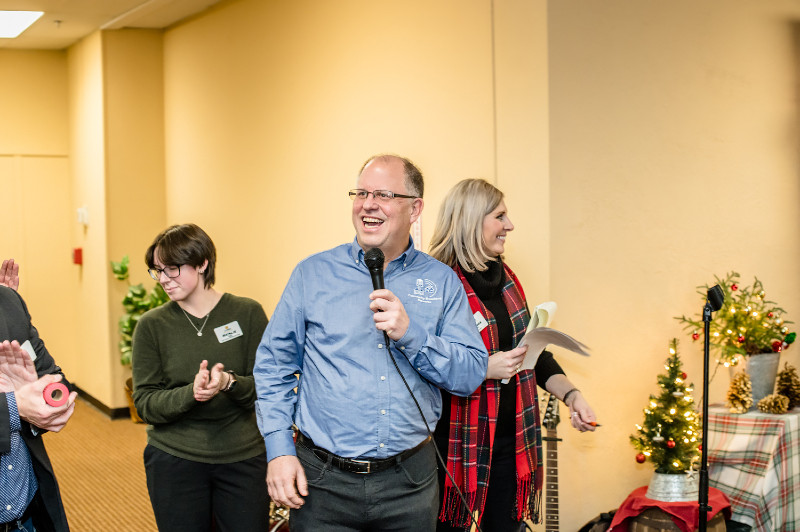 A man in glasses laughs while speaking into a microphone at a holiday event, with others clapping and smiling.