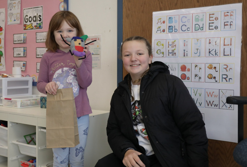 Two girls smile in a classroom; the younger holds a paper bag and a colorful felt craft project.