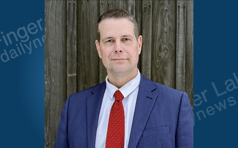 Rob Franklin, a man in a blue suit and red tie, stands against a weathered wooden wall.