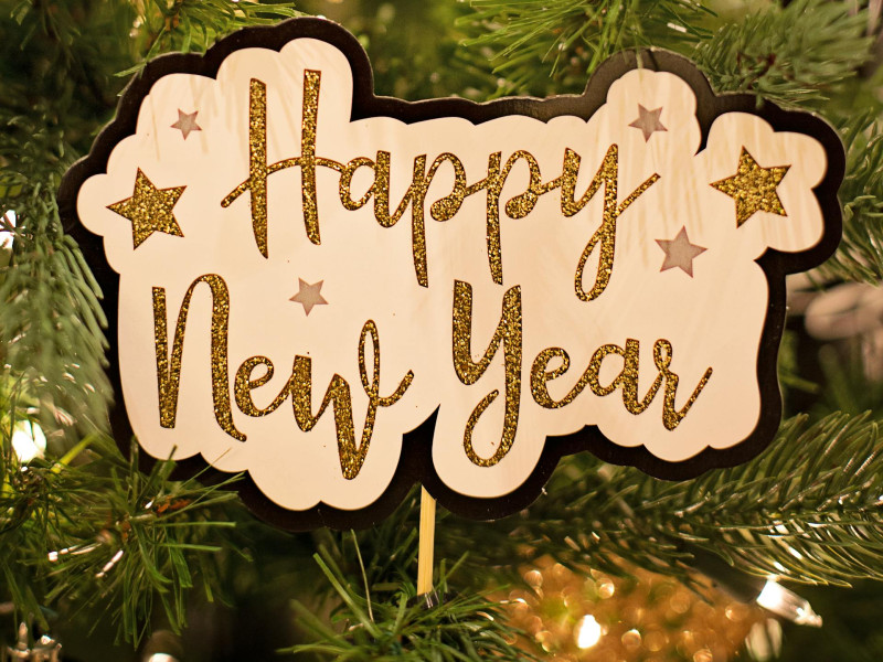 A festive "Happy New Year" sign with gold glitter lettering and stars hangs on a decorated evergreen tree.