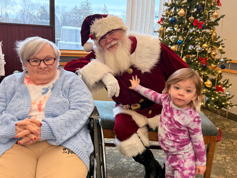 Santa, a child, and an elderly woman in a wheelchair pose by a Christmas tree.