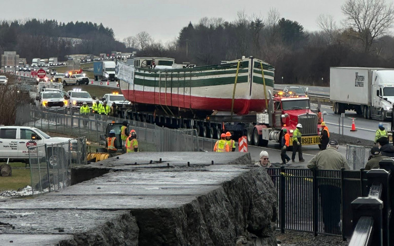 An oversized truck transports the historic Lois McClure canal boat on a highway, surrounded by support vehicles and personnel.