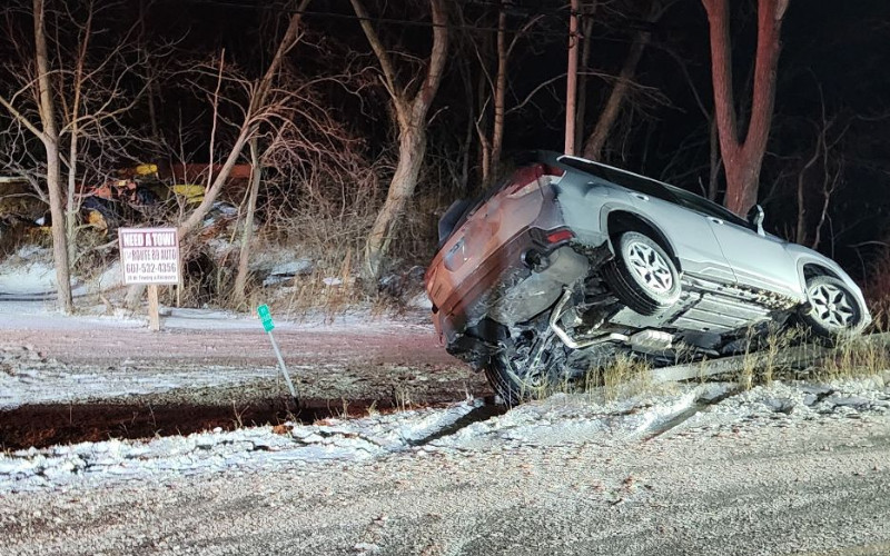 Silver SUV flipped on its side in a snowy ditch at night, near a 'Need a Tow' sign and bare trees.