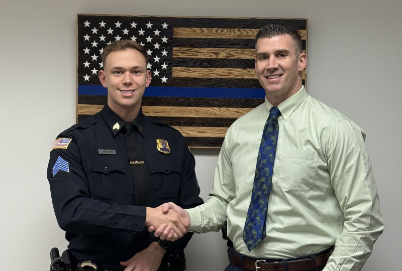 Police officer Tyler Brzezinski shakes hands with another man in front of a wooden Thin Blue Line flag.