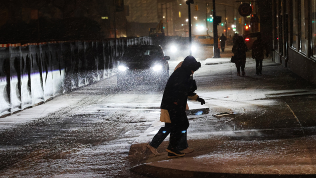 People walk on a snowy street at night as cars drive by with headlights on during a winter storm.