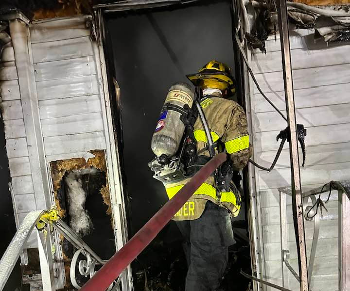 Firefighter in protective gear with hose enters a fire-damaged home.