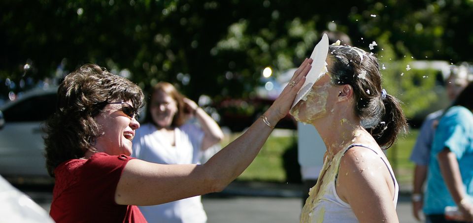 Woman in red shirt throws a pie into another woman's face at an outdoor event, pie splattering.