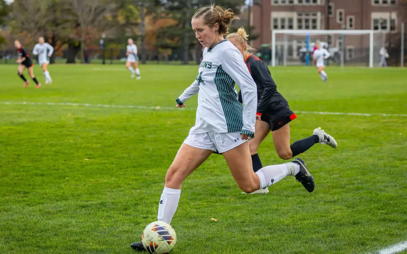 Ruth Hotaling in a white and green soccer uniform kicks a ball on a grassy field during a game.