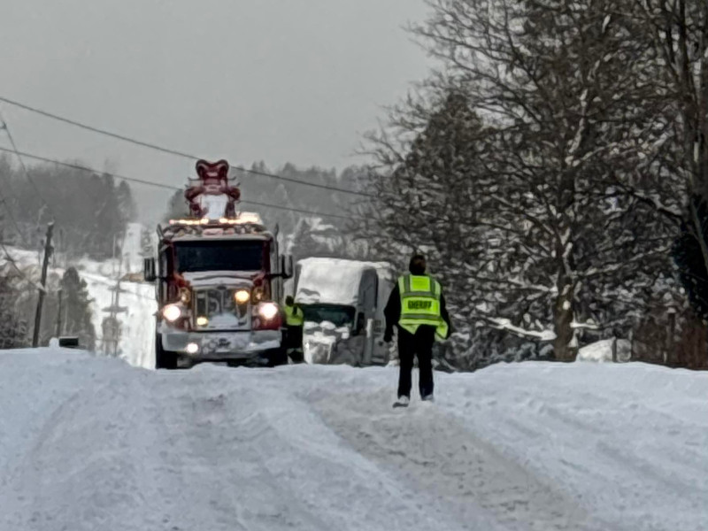 Tow truck pulls a snow-covered vehicle on a snowy road with a sheriff's deputy standing by in Finger Lakes region.