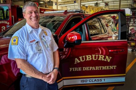 Auburn Assistant Fire Chief Sherman smiles in his uniform in front of a red Auburn Fire Department vehicle.