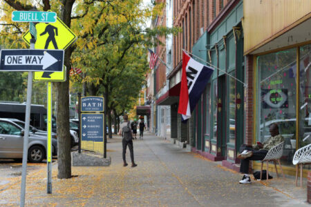 Downtown Bath, NY street scene with storefronts, pedestrians, and street signs including 'Buell St' and a 'Downtown Bath' directory.