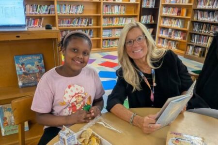 A smiling young girl and an adult woman, likely a 'Book Buddy,' read together in a library.