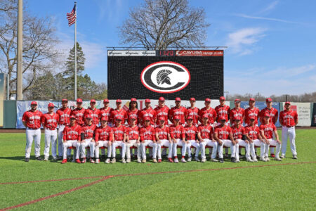 The CCC Spartans baseball team poses for a group photo on their home field with a scoreboard featuring their logo.