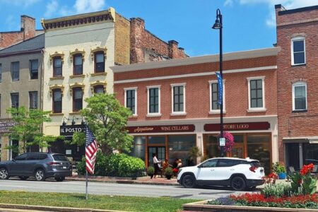Downtown Canandaigua street with historic buildings, shops, cars, and an American flag under a blue sky.
