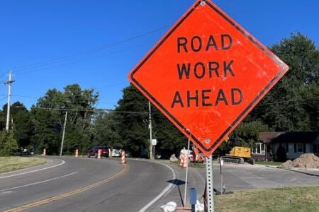 An orange 'ROAD WORK AHEAD' sign stands prominently beside a curving road where construction is underway.