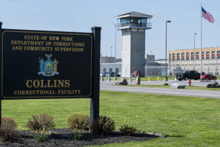 Sign for Collins Correctional Facility with a guard tower and prison buildings in the background.