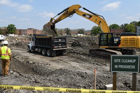 Excavator loads dirt into a dump truck at a cleanup site in Newark, with workers and a 'Site Cleanup in Progress' sign.