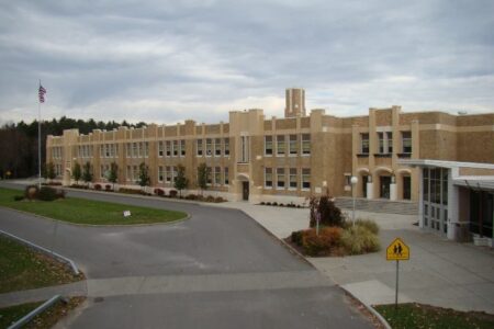 Large brick school building with a driveway, flagpole, and school crossing sign under an overcast sky.