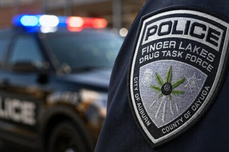 Close-up of a Finger Lakes Drug Task Force patch on a police uniform, with a blurred police car in the background.