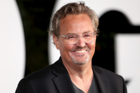 Actor Matthew Perry smiles, wearing glasses and a dark jacket, against a blurred background.